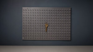 Ein hölzernes Schlüsselbrett mit vielen kleinen Haken, aufgenommen von vorne. Fast alle Haken sind leer, nur an einem hängt noch ein einzelner Messingschlüssel. Der Hintergrund ist dunkles Anthrazit, das Bild ist ruhig und gedämpft.