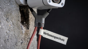 Nahaufnahme einer weissen Überwachungskamera an einer Betonwand. Ihr Kabel teilt sich an einem Verteiler: Ein Strang führt zurück in die Wand, ein zweiter, rotbrauner Strang führt weg und trägt ein Kabeletikett mit der Aufschrift „Verfassungsschutz". Das Bild steht für den geplanten Zugriff des deutschen Verfassungsschutzes auf private Videoanlagen.